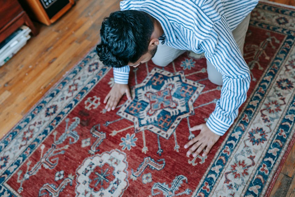 A close-up of intricately patterned Persian-style rugs with detailed floral and geometric motifs in shades of blue, beige, and gold, layered on a wooden floor. The rugs have a soft, plush texture with
