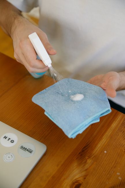 A person wearing light green rubber gloves is kneeling on a shiny white tiled floor, using a green and dark blue cleaning sponge to scrub the surface. The room features wooden furniture and shelves in
