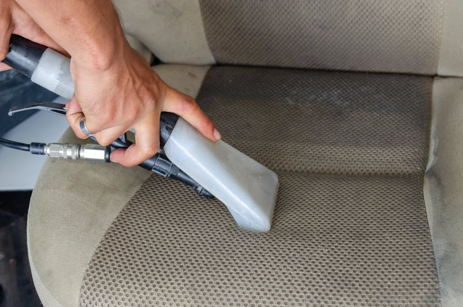 A close-up view of a person's hand operating a handheld upholstery cleaning machine on a beige fabric armchair in a room illuminated by natural light. The machine's nozzle is pressed firmly against the armrest, which features a textured, patterned surface, with some visible stains and dirt. In the background, part of the cushioned backrest and seat can be seen, both showing signs of usage. The scene highlights detailed surface cleaning and sanitisation processes provided by Carpet Cleaners SW6, demonstrating professional deep cleaning of upholstery fabrics to restore cleanliness and hygiene in a residential setting, referencing Stamford Bridge upholstery cleaning after match day stains, SW6.